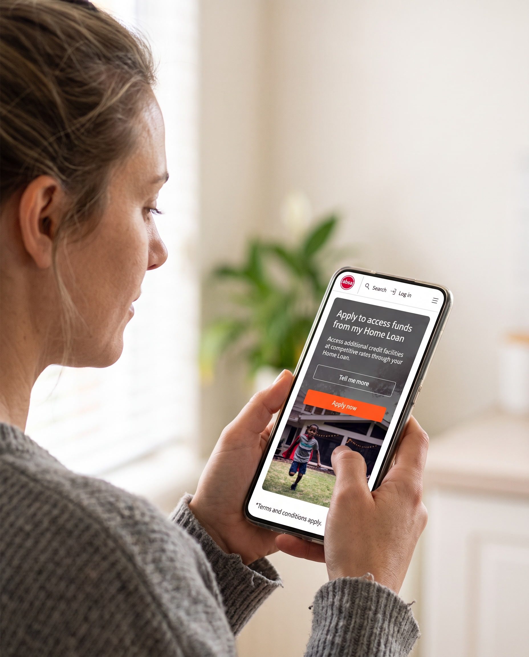Close over a woman’s shoulder as she reads Absa’s mobile home loans page on her phone in a sunlit kitchen: the headline Apply to access funds from my Home Loan, a paragraph about additional credit facilities at competitive rates, a Tell me more button and an orange Apply now button.
