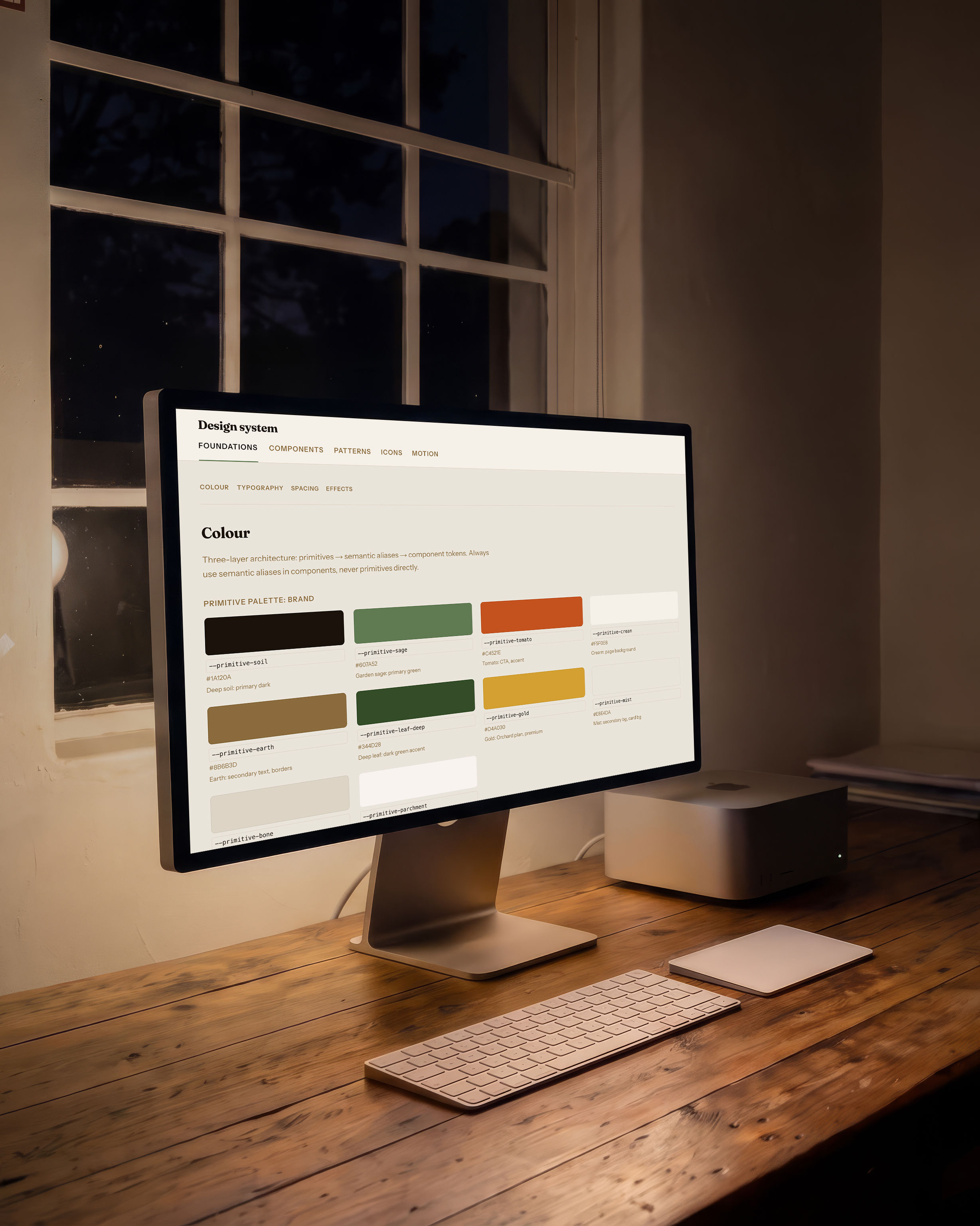 A large display on a timber desk in a studio at night, showing the GardenHarvestAI design system: the Foundations tab open on Colour, a note that the architecture runs from primitives through semantic aliases to component tokens, and a grid of brand colour swatches each labelled with its token name, its hex value and what it is for.