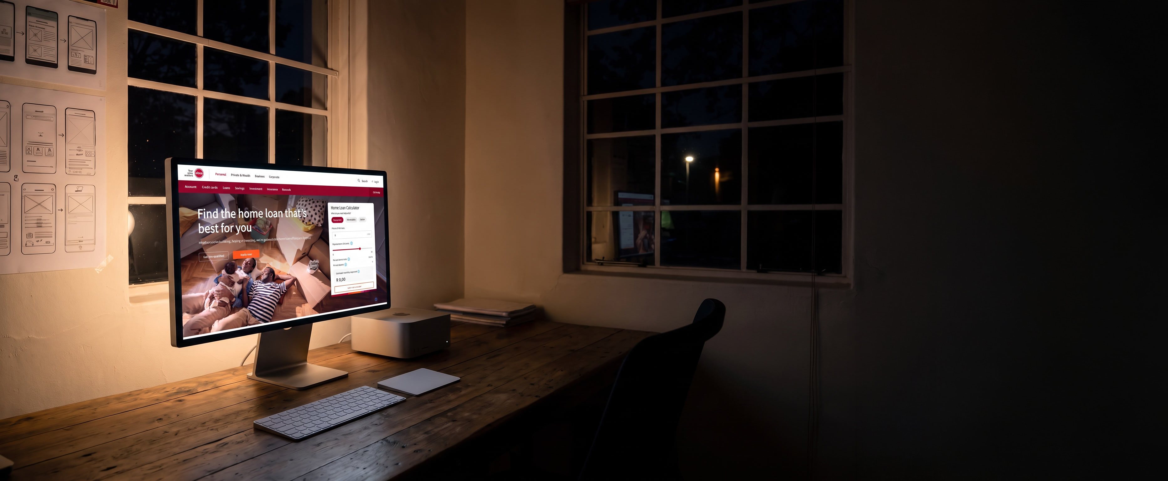 A designer's studio at night, lit only by the display on the timber desk. Absa's home loans page is open on it, the headline Find the home loan that's best for you over a photograph of a family, with a rates panel beside it. A keyboard and trackpad sit in front, a small desktop computer behind, printed wireframes are pinned to the wall on the left, and two tall paned windows look out on the dark.