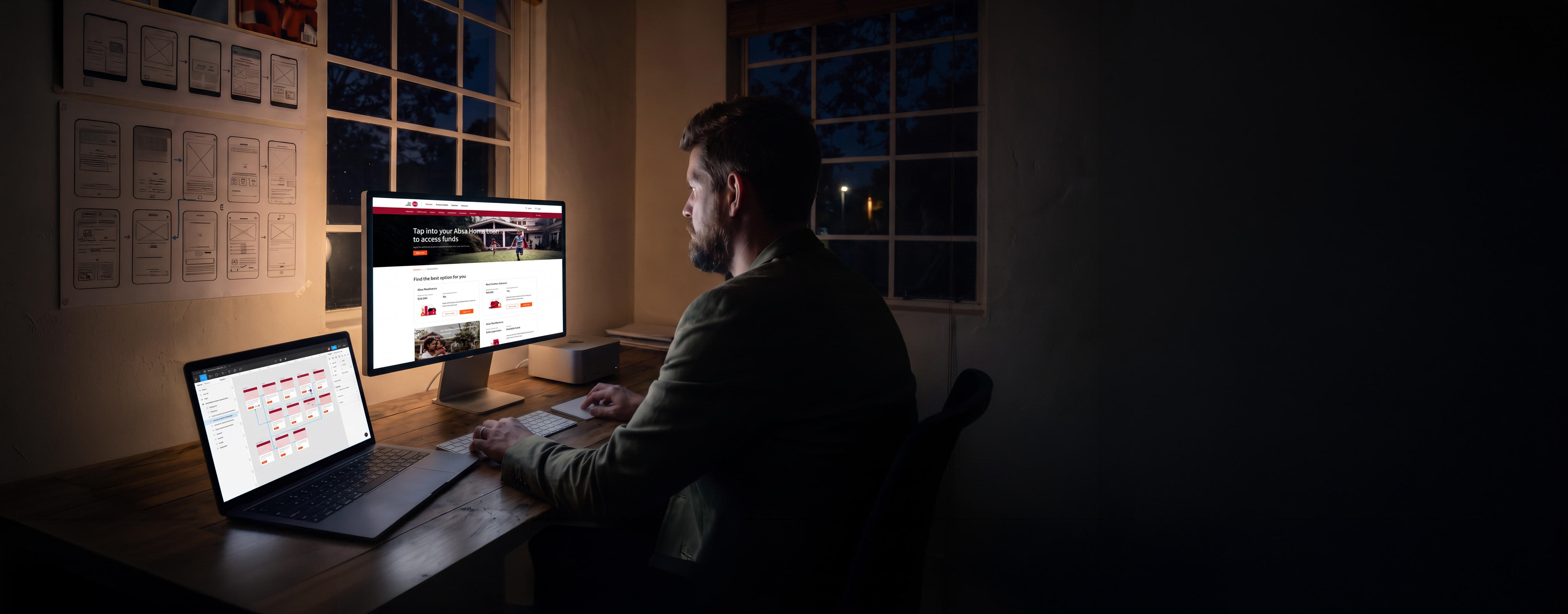 Will Derman seen from behind at his desk in the studio at night, lit by his screens: Absa's home loans page on the display, a design file open on the laptop beside it, and a wall of printed wireframes pinned up to his left.