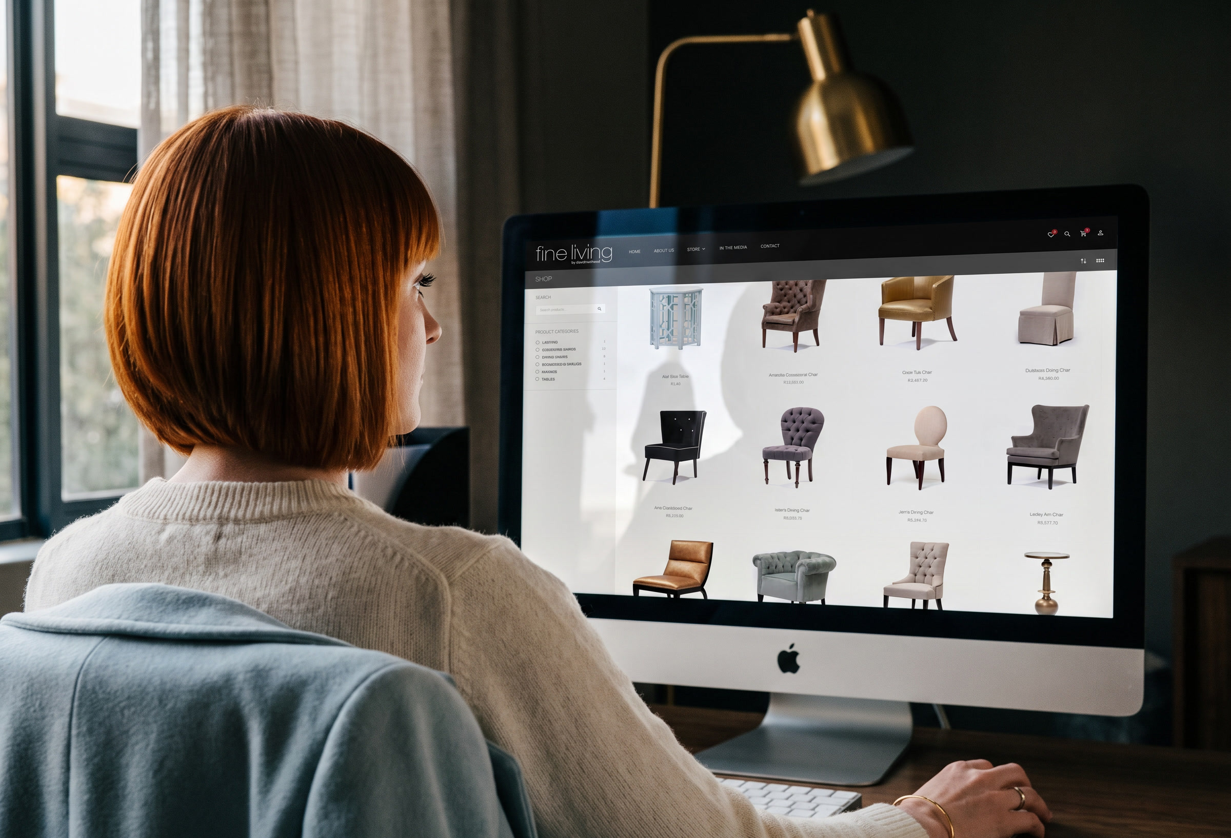 A woman at a desktop computer in a dark, warm room, the Fine Living shop open on screen showing the chair range in a grid.