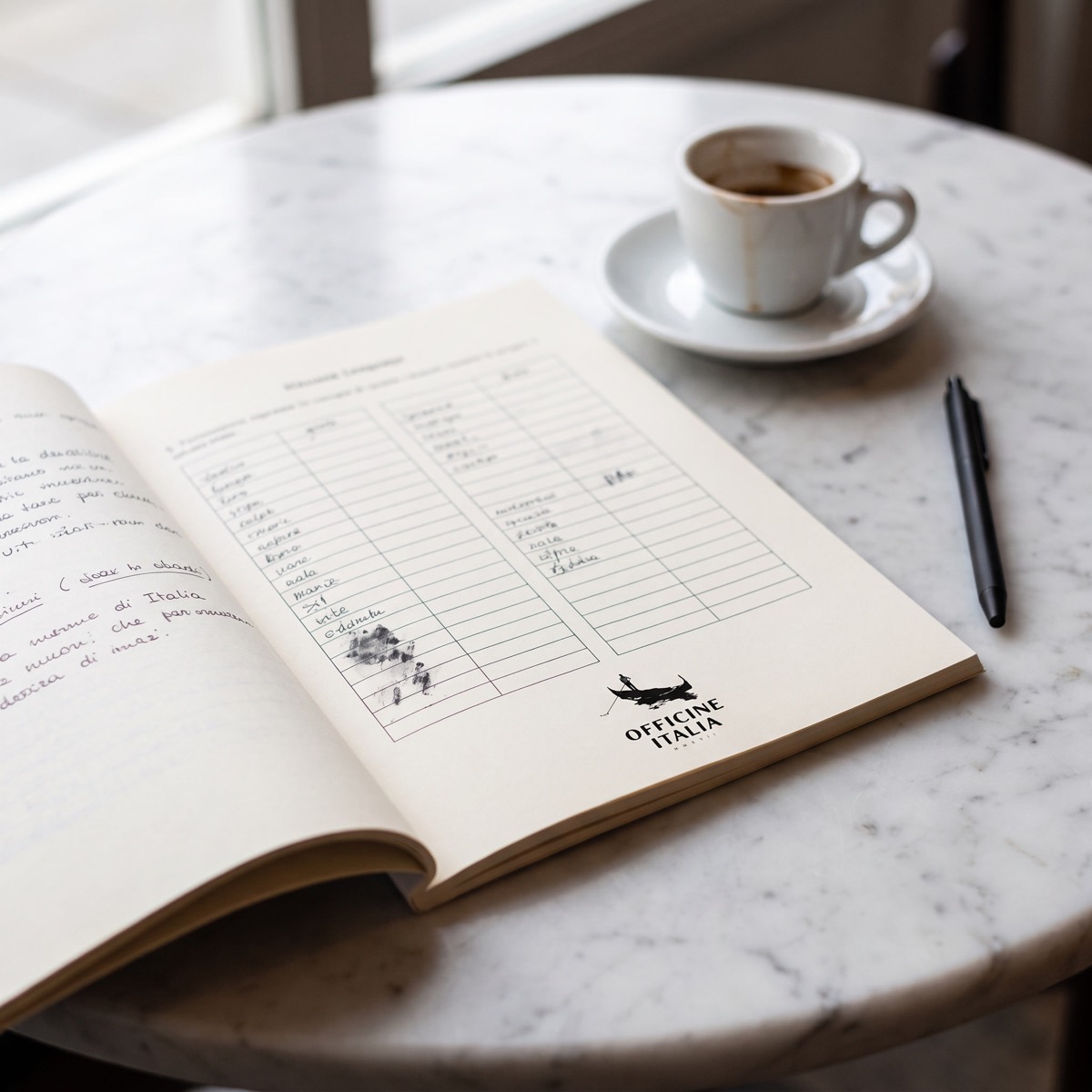 An Italian language workbook open on a marble café table, the Officine Italia mark small at the foot of the page.