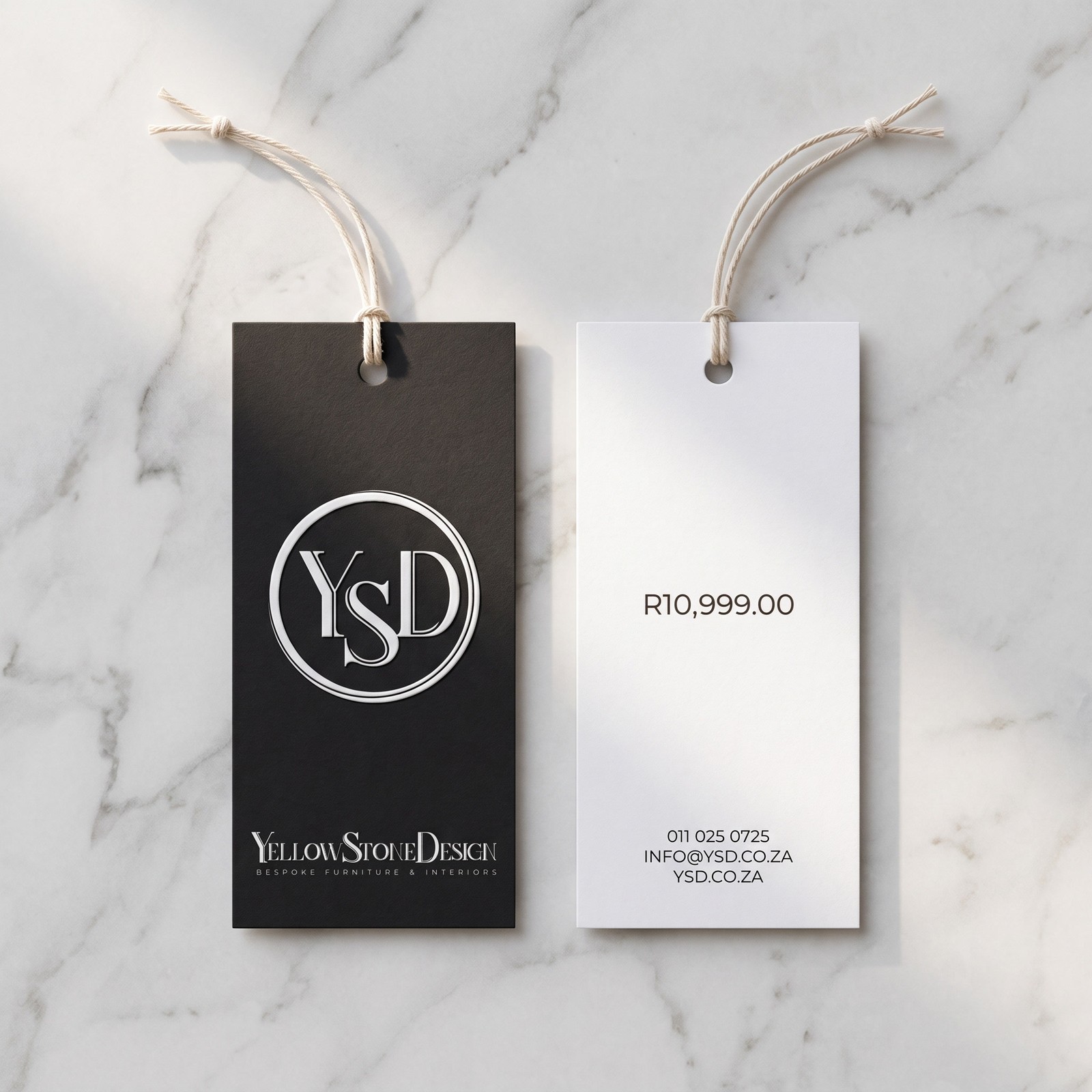 Two swing tags on pale marble: a black one face up carrying the embossed YSD monogram and the wordmark, and a white one face down carrying the price and the contact details.